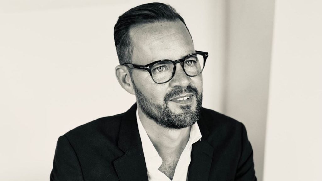 Black and white image. Man in black suit and white shirt, black glasses, plain background.