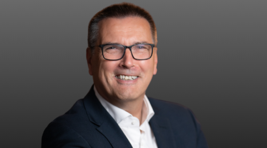 man in suit with white shirt, black glasses looking at camera smiling, grey background